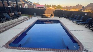 A rectangular swimming pool with a brick border, surrounded by lounge chairs and a wooden table.