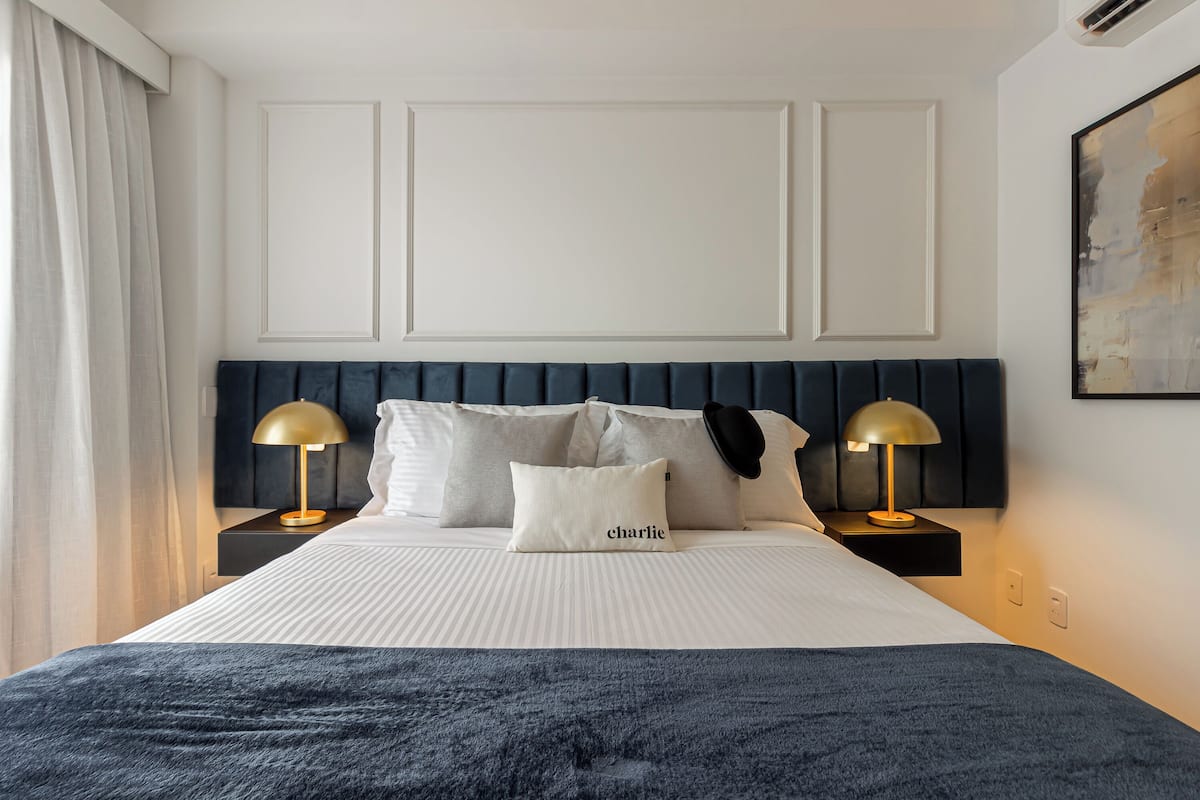 Una cama bien hecha con un cabecero oscuro, dos lámparas de noche y una almohada con el nombre 'charlie'.