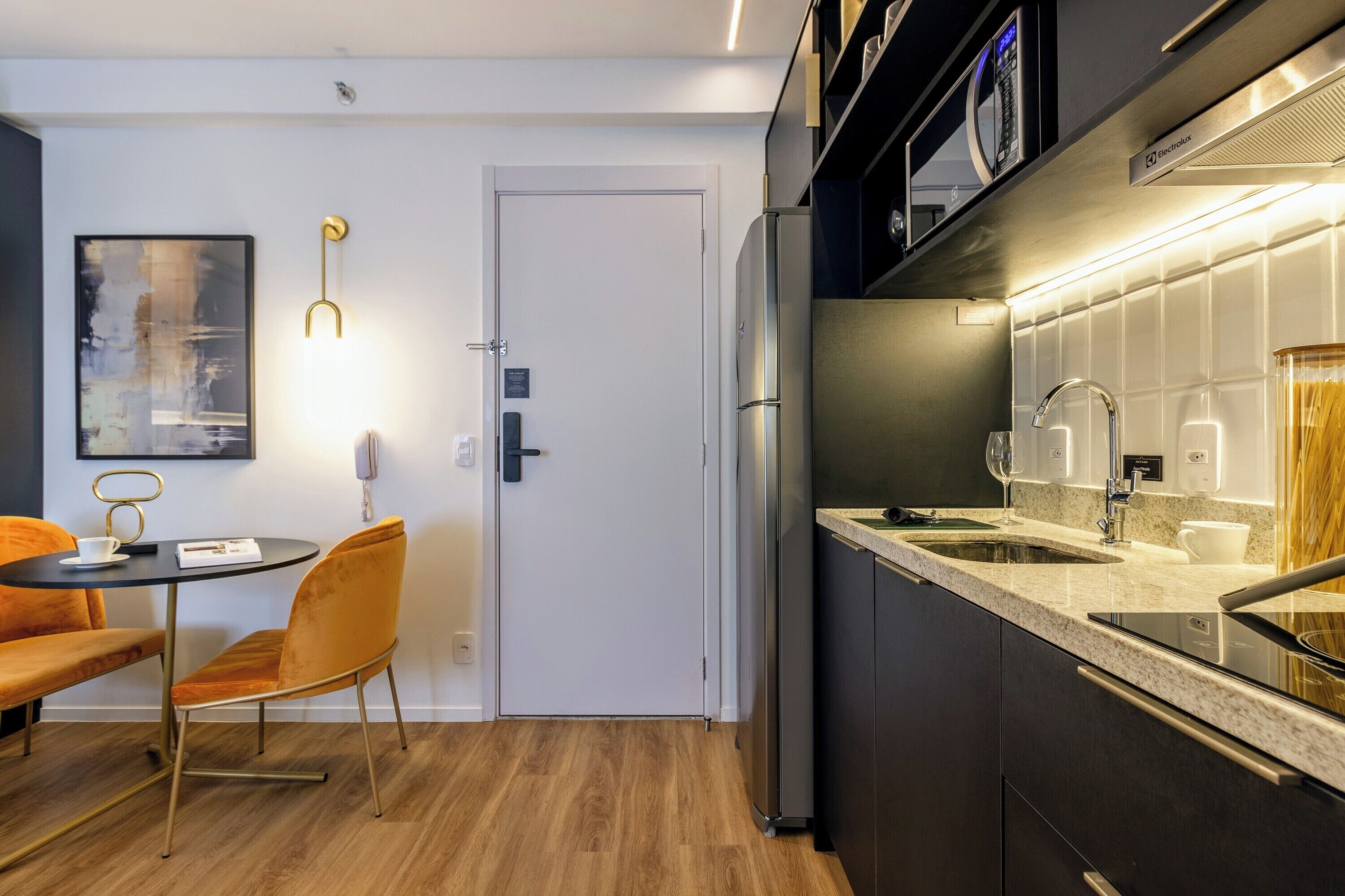 A modern kitchen with a stainless steel refrigerator, a built-in oven, and a sink. There is a dining area with a round table and two chairs.
