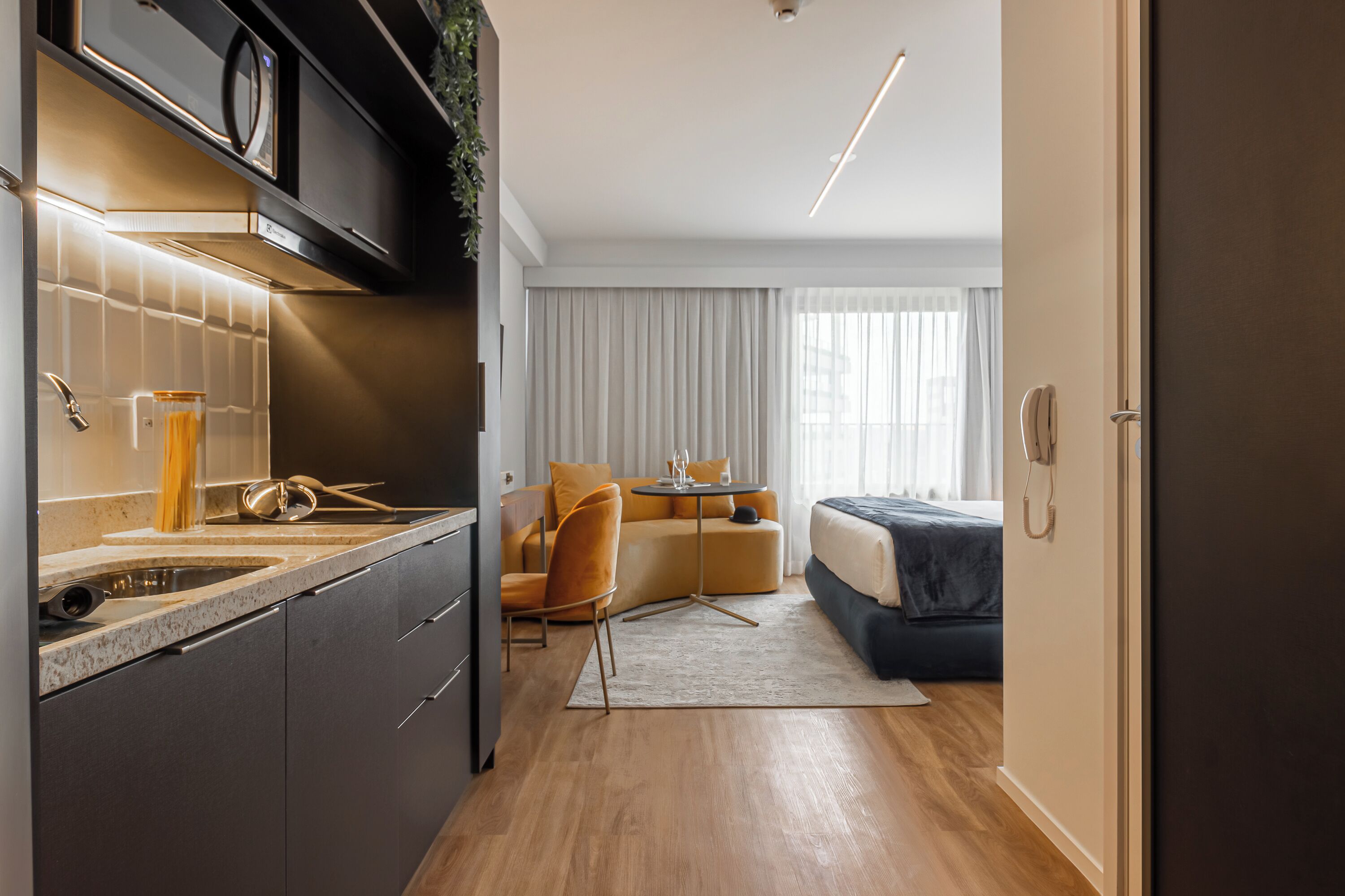 A modern kitchen with a built-in oven, a dining area with a round table, a sofa, and a bed in the adjacent room.