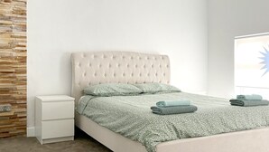 A bedroom with a tufted headboard, a white nightstand, a bed with a floral-patterned bedspread, and two rolled towels.