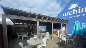 An outdoor patio area with white plastic chairs and tables, a covered seating area, and a sign for 'Urchin Bar & Adventures.'