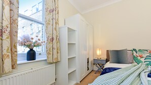A bedroom with a window, a radiator, a bed with a blue and green patterned bedspread, a nightstand with a lamp, and a wardrobe. - Rathbone Exclusive - ES1 (London)