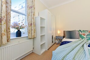 Ein Schlafzimmer mit Fenster, Heizkörper, einem Bett mit blau-grün gemustertem Überwurf, einem Nachttisch mit Lampe und einem Kleiderschrank.