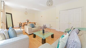 A living room with a sofa, a glass coffee table, a chandelier, and a dining area with a table and chairs. - Rathbone Exclusive - ES1 (London)
