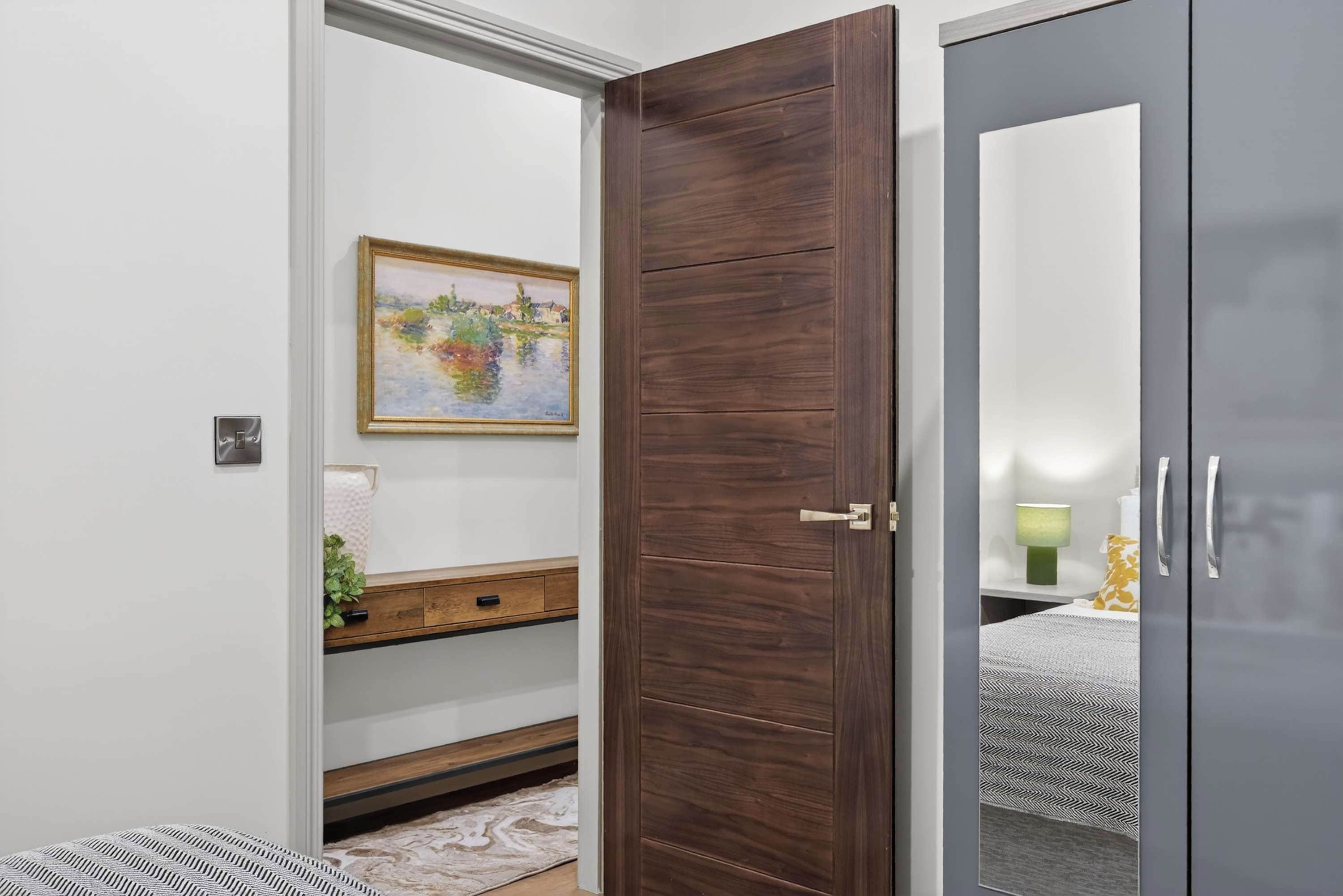 Una habitación con puerta de madera, un cuadro en la pared, una mesita de noche con lámpara y una cama con colcha estampada.