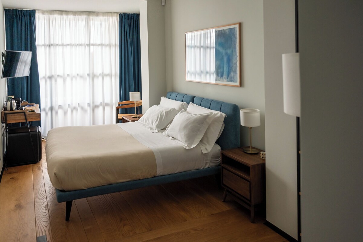 Habitación de hotel con cama, escritorio, silla, lámpara y un cuadro en la pared.