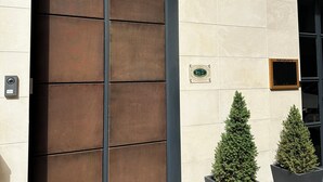 A Club 64 building entrance with a large metal door, a sign indicating a restaurant, and two potted evergreen trees. - Club64  (Andria)