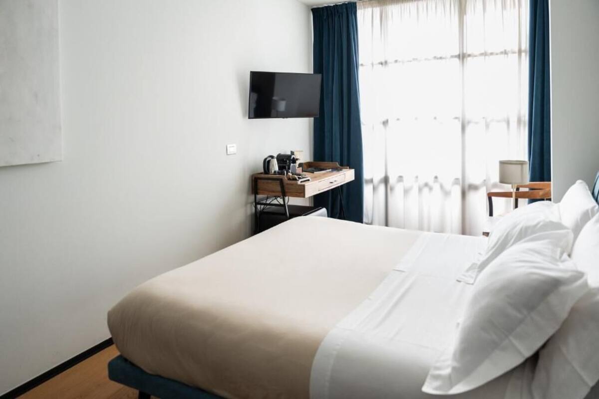 A hotel room with a bed, a desk with a flat-screen TV, and a window with curtains.