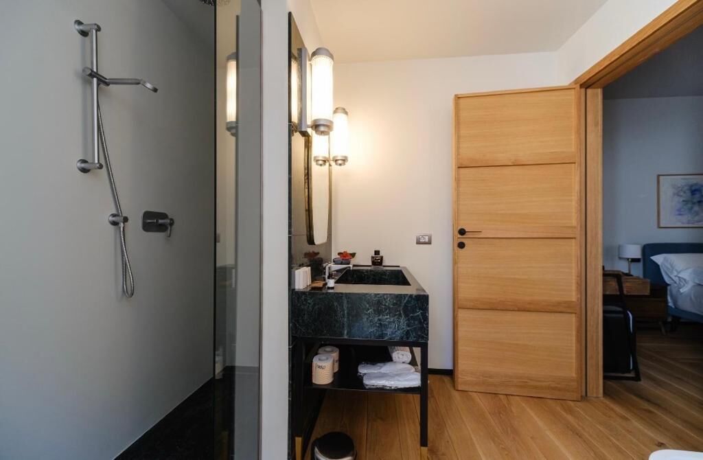 A compact bathroom with a shower, a sink with a marble countertop, and a wooden door leading to a bedroom.