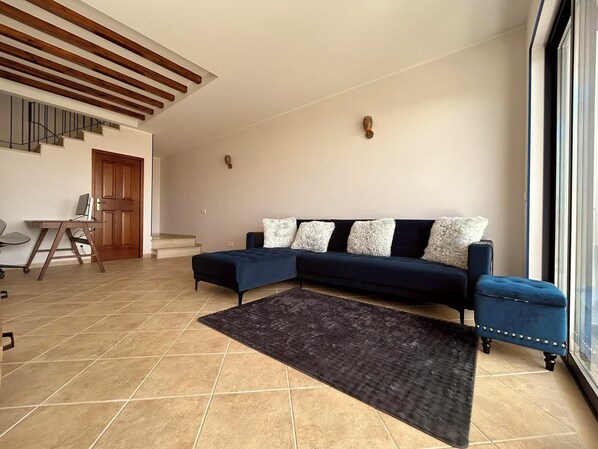 A modern living room with a sectional sofa, a dark rug, a desk with a chair, and a staircase.