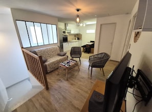 Un salon avec un canapé, une table basse, deux fauteuils, un téléviseur et, au fond, une cuisine.