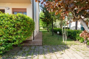 Ein gepflasterter Weg führt zu einem Gebäude, begleitet von einer hohen Hecke auf der einen Seite und einem Baum mit Herbstlaub auf der anderen.