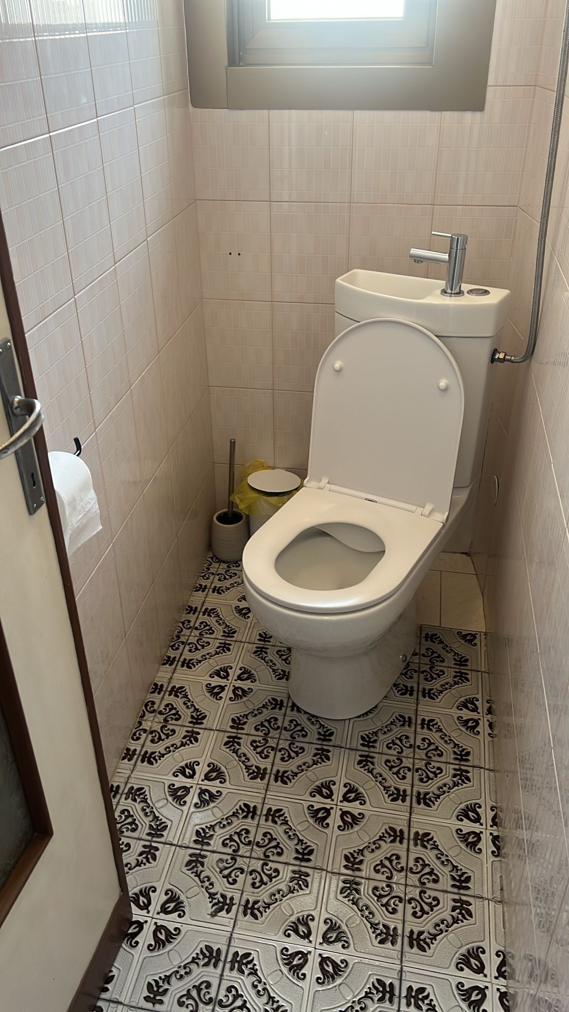 A small bathroom with a toilet, a bidet, a toilet paper holder, and a window above the toilet.