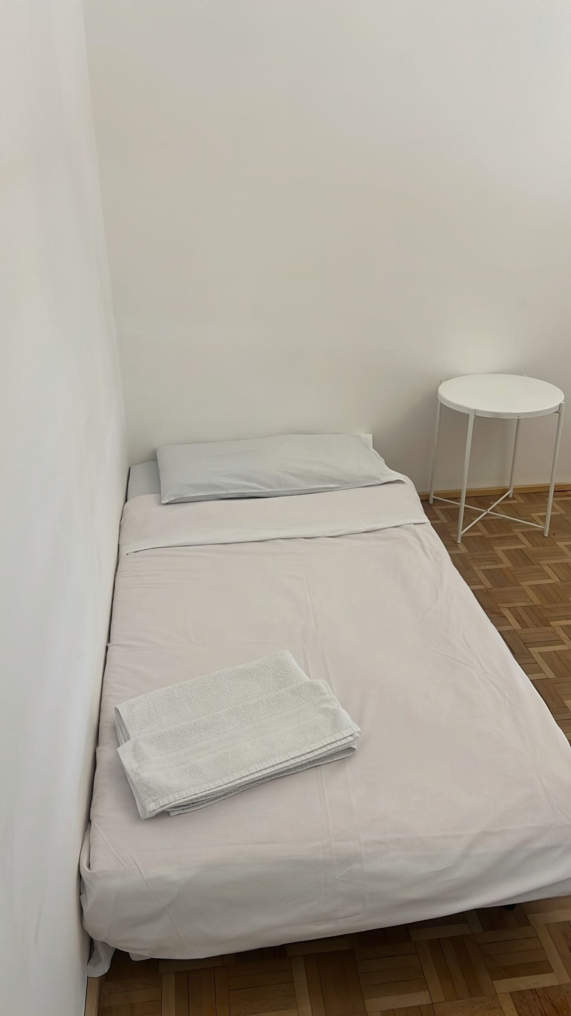 A small, single-bed room with a white bedspread, a folded towel, and a small white table.