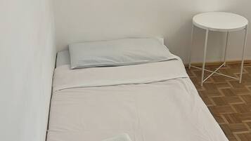 A small, single-bed room with a white bedspread, a folded towel, and a small white table.