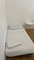 Piccola camera con un letto singolo, copripiumino bianco, un asciugamano piegato e un tavolino bianco.