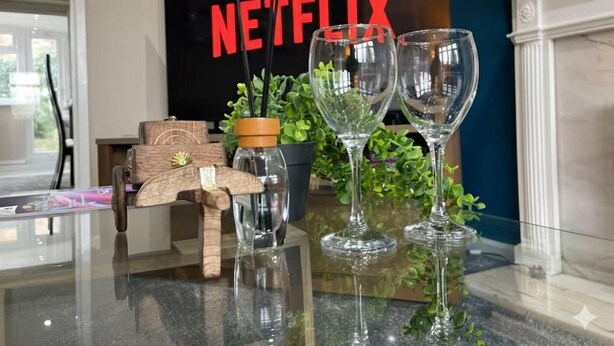 A room with a television displaying 'NETFLIX,' a glass table with wine glasses and a bottle, and a wooden object with a tag. - House Sleeps 6 With Garden and Parking (Leeds)