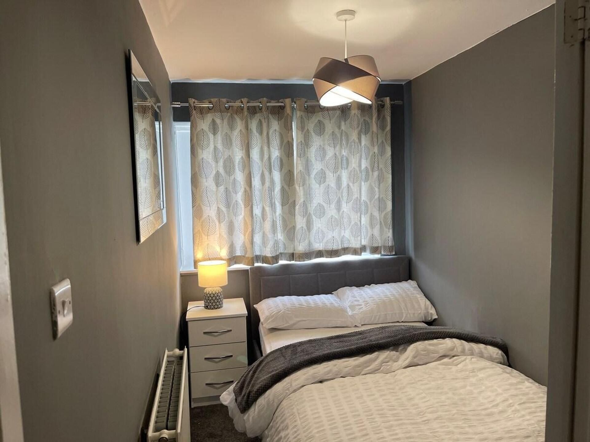 Un dormitorio con cama, una mesita de noche con lámpara, un radiador y una ventana con cortinas.