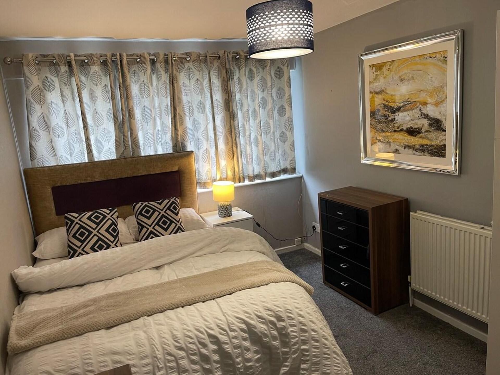 A bedroom with a bed, a nightstand, a lamp, a dresser, a radiator, and a painting on the wall.