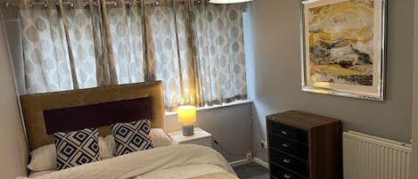 A bedroom with a bed, a nightstand, a lamp, a dresser, a radiator, and a painting on the wall.