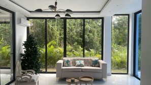 Ein modernes Wohnzimmer mit einer Couch, zwei kleinen Tischen, einem Zierstuhl und einem großen Fenster mit Blick auf Grünflächen.