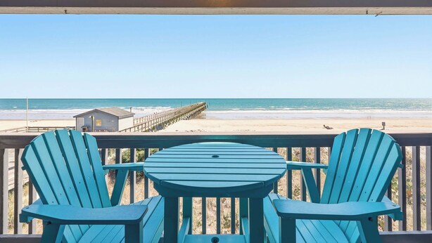 A balcony with two chairs and a table, overlooking a beach with a pier.