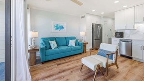A modern living room with a teal sofa, a white armchair, a ceiling fan, and a kitchen area with white cabinets and stainless steel appliances.