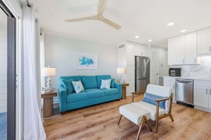 A modern living room with a teal sofa, a white armchair, a ceiling fan, and a kitchen area with white cabinets and stainless steel appliances. - Tide Time Oceanfront at Sea Cabin w Pool (North Myrtle Beach)