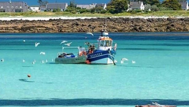 A coastal scene with a boat, clear turquoise water, and a rocky shoreline with buildings. - Ty Argoat -  Your haven between land and sea (Plouarzel)