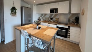 Eine moderne Küche mit weißen Schränken, einem Edelstahlkühlschrank, einer Mikrowelle und einem Geschirrspüler. Ein Holztisch mit zwei Stühlen steht bereit.