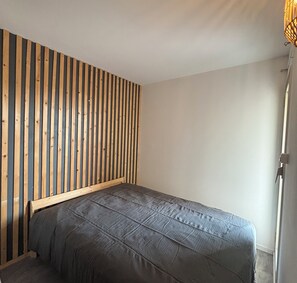 Ein Schlafzimmer mit einem querverlegten Holzkopfteil, einem Einzelbett mit grauer Bettdecke und einer schlichten weißen Wand.