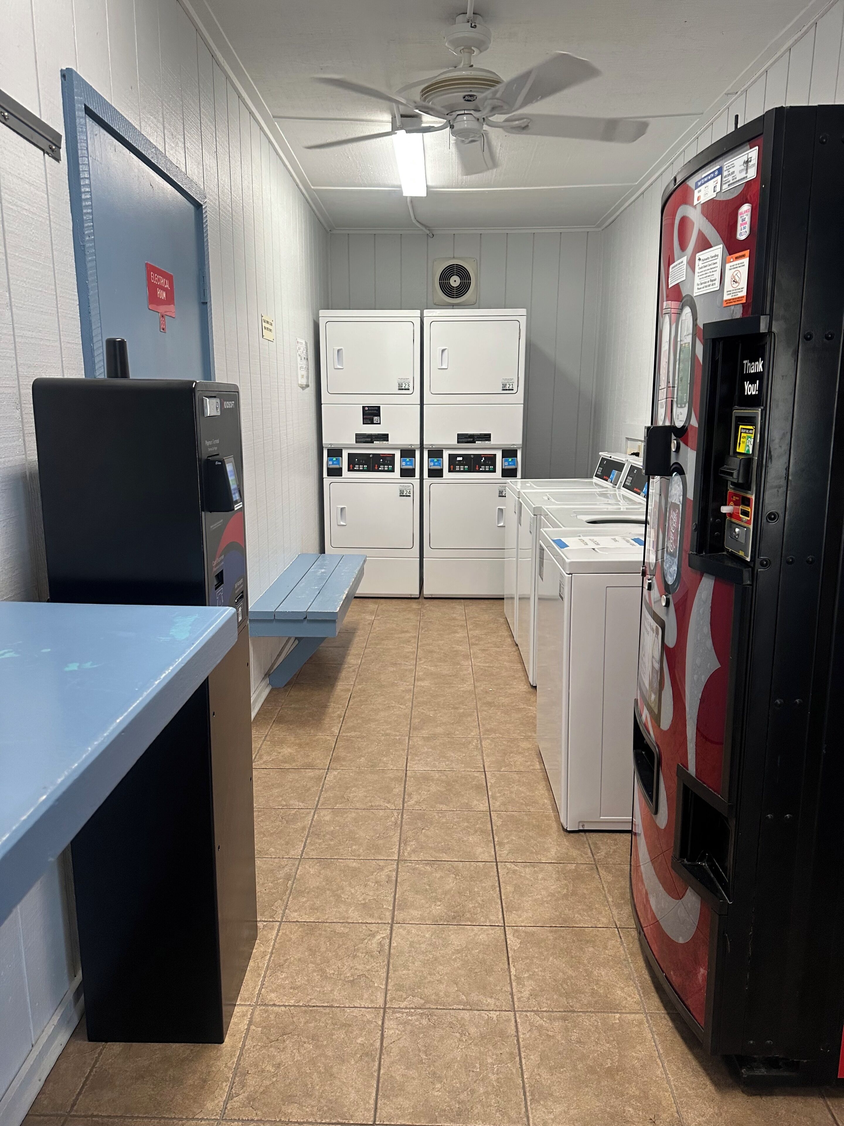 Uma lavanderia com uma bancada azul, uma máquina de vendas automática e secadoras empilhadas.