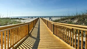 Een houten looppad dat naar een strand leidt, met zandduinen en een helderblauwe lucht.
