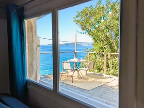 A room with a large window overlooking a balcony with a table and chairs, a view of the sea, and a tree. - Rustic house with amazing sea view (Le Pradet)
