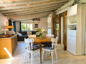 A dining area with a wooden table, white chairs, a vase with flowers, a sofa, and a kitchen area with a refrigerator. - Rustic house with amazing sea view (Le Pradet)