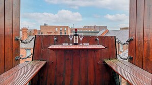 Un balcón de madera con una botella de vino y copas, con vistas a un paisaje urbano.