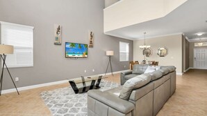 A spacious living room with a large gray sectional sofa, a glass coffee table, and a painting of a beach scene on the wall. - Spacious Family Home w Pool, Hot Tub & BBQ (Kissimmee)