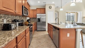 A modern kitchen with wooden cabinets, granite countertops, stainless steel appliances, and a large island with a sink. - Spacious Family Home w Pool, Hot Tub & BBQ (Kissimmee)