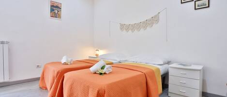 A hotel room with two beds, a bedside table, a nightstand, and a wall-mounted macrame piece.