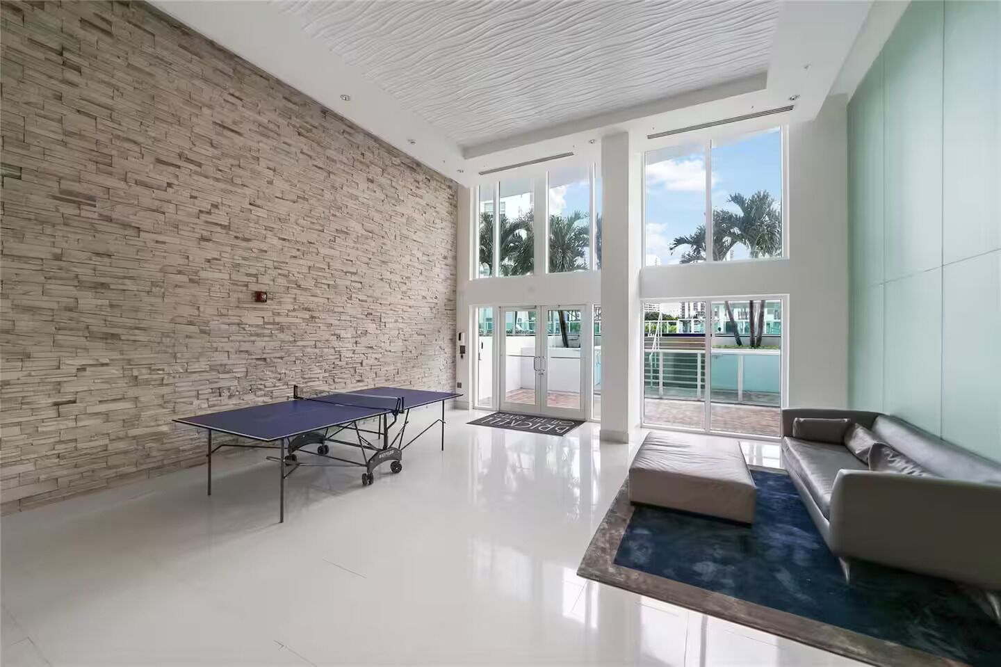 A spacious room with a ping pong table, a sofa, and a view of a pool through large windows.