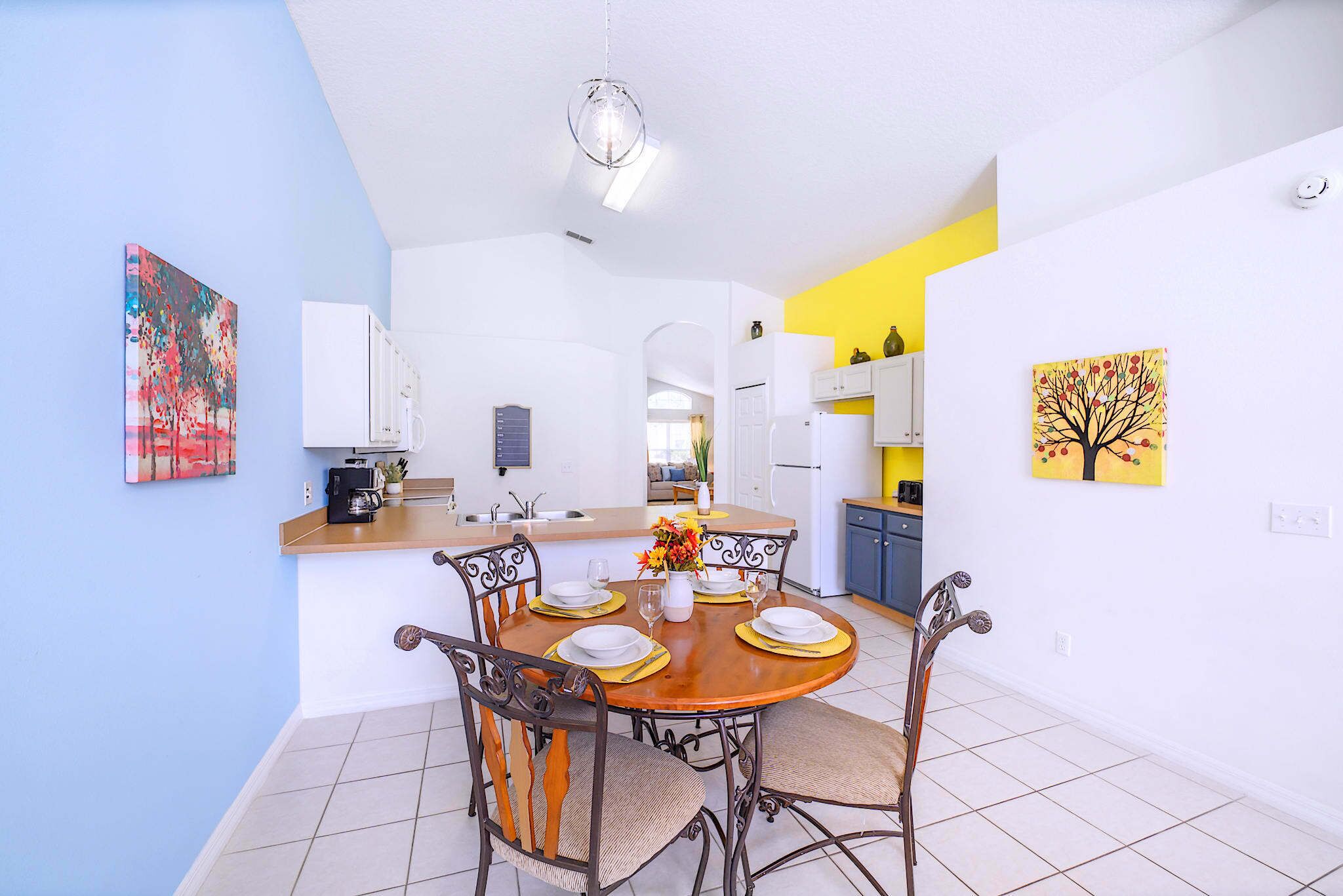 A dining area with a round table set for four, surrounded by chairs. There is a kitchen in the background with white cabinets and a yellow accent wall.