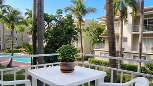 Un balcón con una mesa y sillas blancas, con vista a una piscina y palmeras.