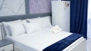 Habitación de hotel con cama, almohadas blancas, manta azul, mesita de noche blanca, armario blanco y ventana con cortinas.