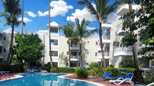 Una piscina con sillas de descanso, rodeada de palmeras y un edificio de varios pisos con balcones.