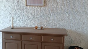 A room with a textured wall, a framed artwork, a brown cabinet with drawers, a small stool, and a candle holder. - Lovely apartment 2 steps from the beach and all amenities! (SAINT CYPRIEN)