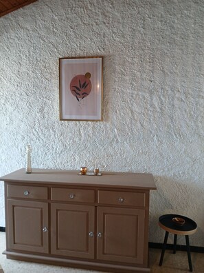 Ein Zimmer mit strukturierter Wand, einem gerahmten Kunstwerk, einer braunen Kommode mit Schubladen, einem kleinen Hocker und einem Kerzenhalter.