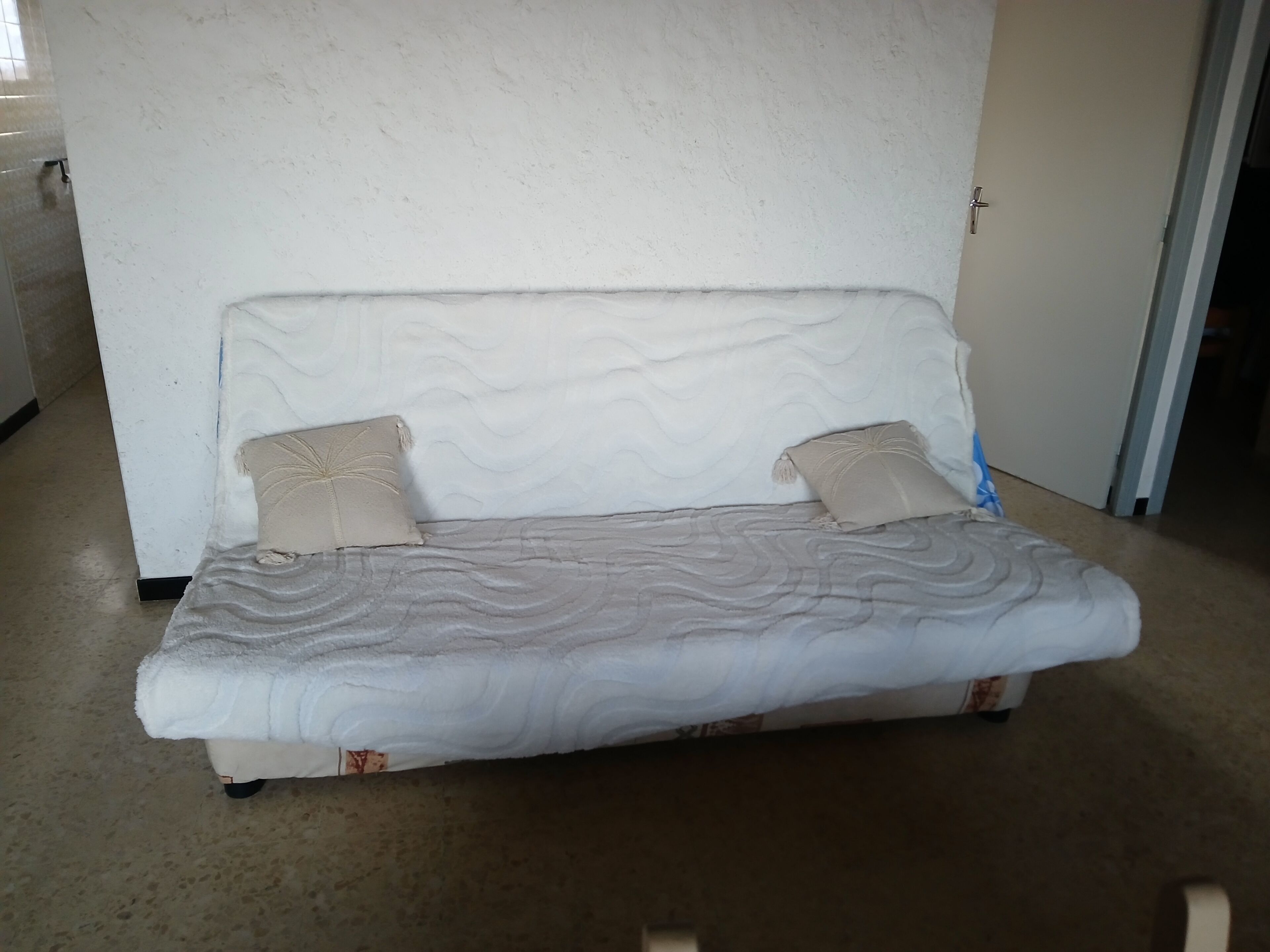 A single bed with a textured mattress and two pillows, positioned against a plain wall with a door to the right.