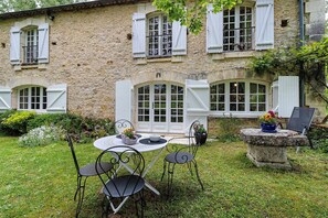 A stone building with white shutters, a round table with chairs, and a small garden area.
