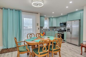 A modern kitchen with teal cabinets, a stainless steel refrigerator, and a dining area with wooden chairs and a table. - Sunny Crest Stay – NEW 2BR, Near Beach (Wildwood)